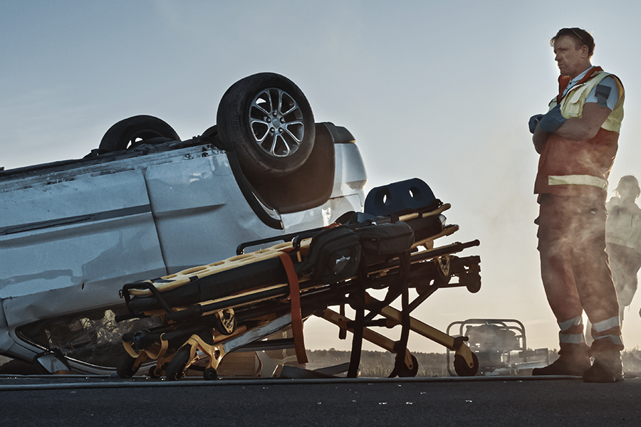 A paramedic standing next to an ambulance gurney and an overturned car.