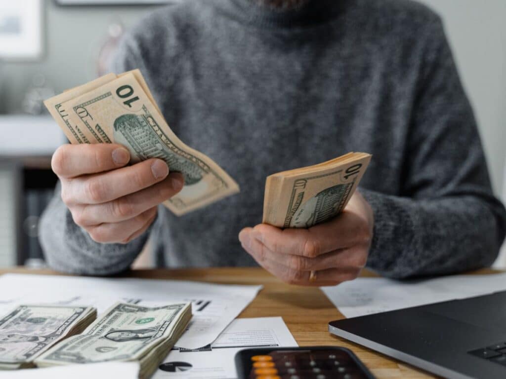 A man in a grey sweater counting money at his desk.