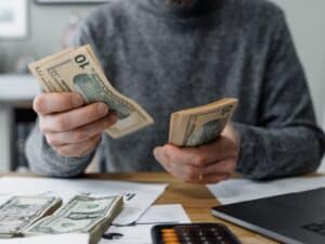 A man in a grey sweater counting money at his desk.