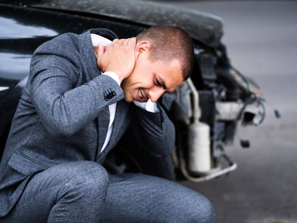 A man holding his neck and grimacing in pain in front of his car that was involved in a collision.