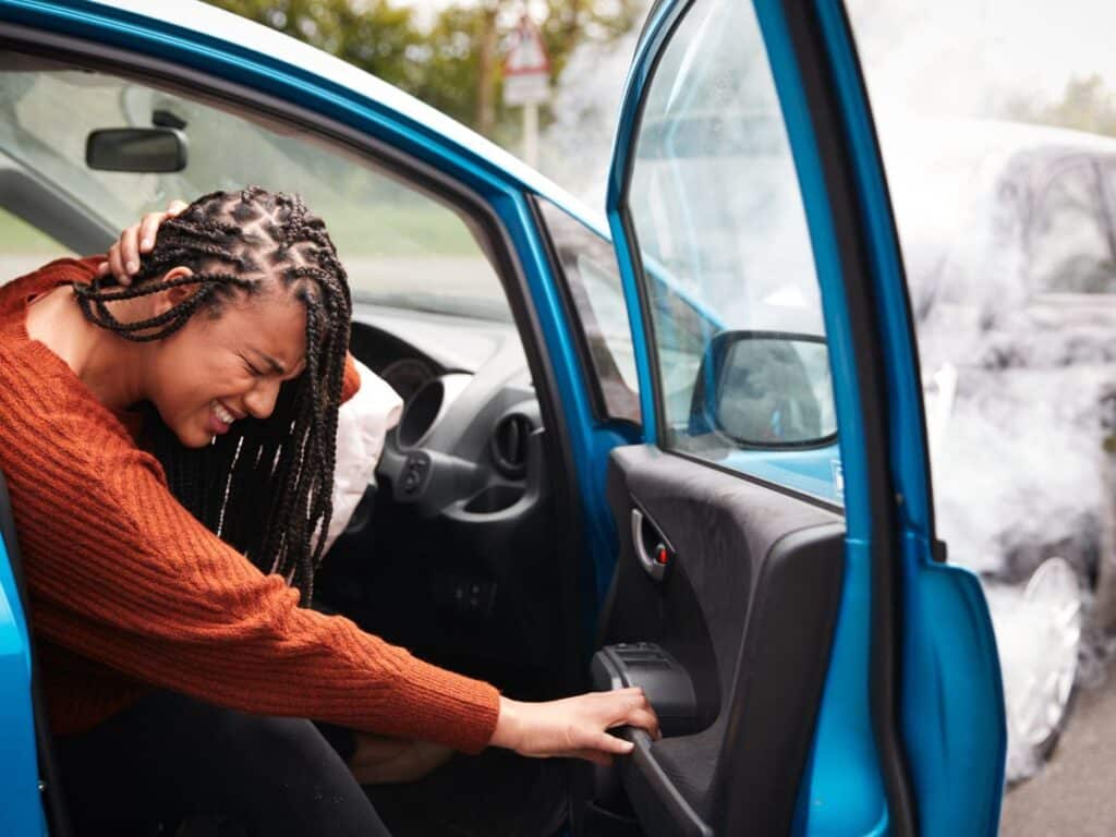 A woman in an orange shirt stepping out of a car and grimacing in pain after a car accident.