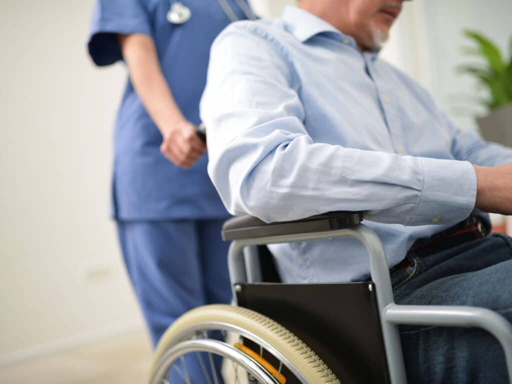 A man in a blue dress shirt sitting in a wheelchair while being escorted out of the hospital by a nurse.
