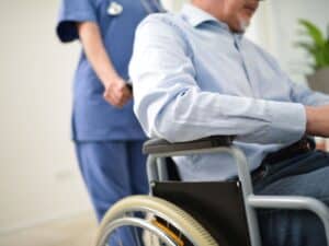 A man in a blue dress shirt sitting in a wheelchair while being escorted out of the hospital by a nurse.