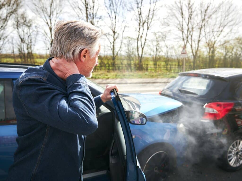 An older man holding his neck and looking at the scene of a two-car collision.