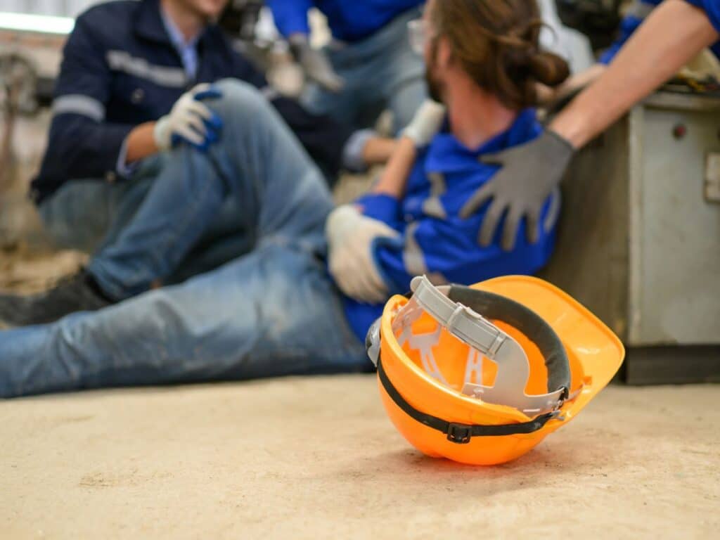 An injured construction worker holding his harm in pain with his yellow hard hat next to him.