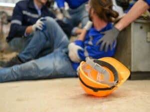An injured construction worker holding his harm in pain with his yellow hard hat next to him.