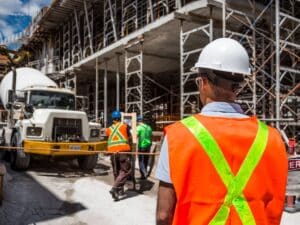 A construction worker wearing an orange and yellow vest and hard hat looking at a construction site.