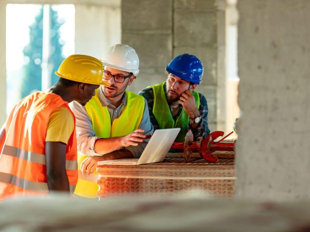 A group of three construction workers in full gear standing around a table discussing plans.