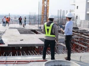 Two construction workers wearing hard hats looking out at a construction site.