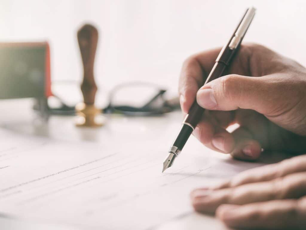 A man's hand signing a document with a gavel in the background.