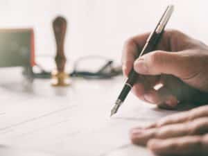 A man's hand signing a document with a gavel in the background.