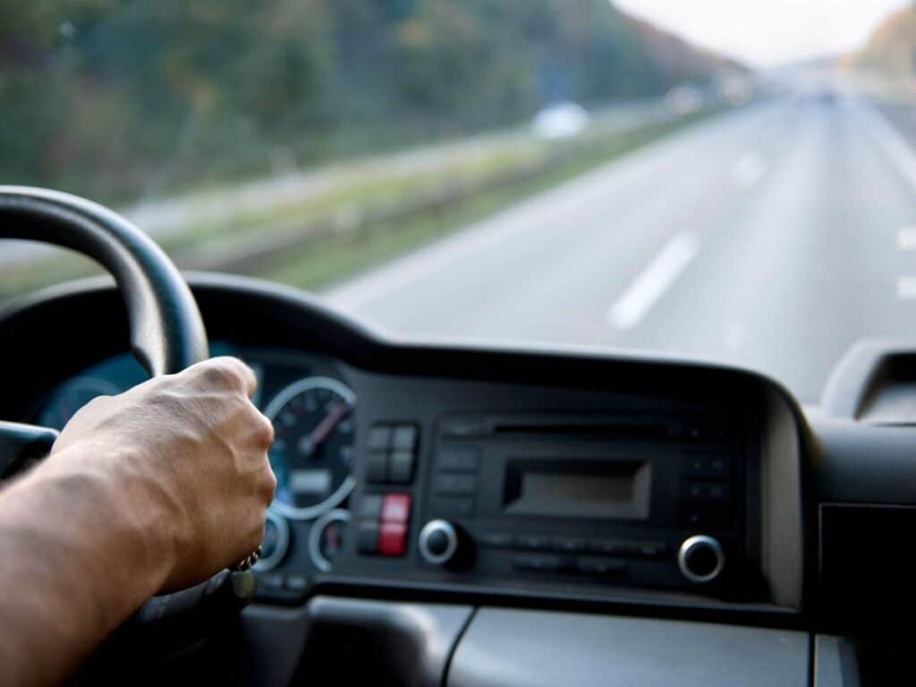 A man's hand gripping the steering wheel of a car as he drives on the highway.