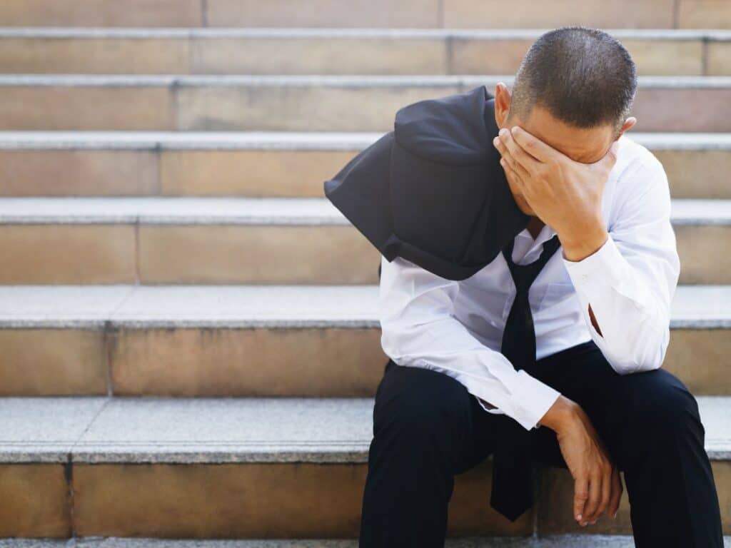 A man sitting on stairs with his head in his hands and a jacket hanging over his shoulder.